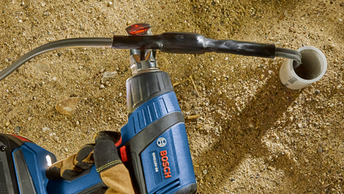 Bosch GHG18V-50 heat gun being used on a wire connection, with a glove, on a surface covered in pebbles. A white PVC pipe is in the background.