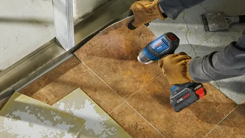 Hands with gloves are using a Bosch GGH18V-50 heat gun, and a Core18V battery pack on a tiled surface.