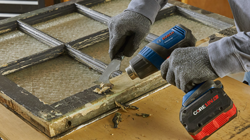 Person using Bosch GHG18V-50 heat gun and a scraper to remove paint from a window frame, with a CORE18V 8Ah battery attached.