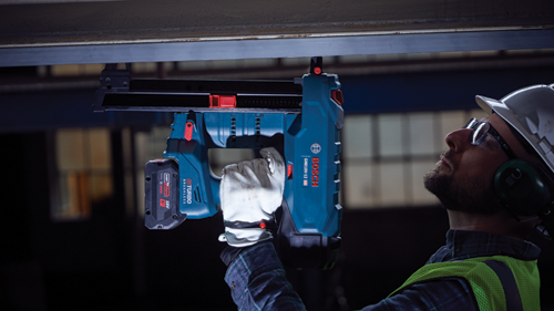 Person wearing safety glasses, ear protection, and a hard hat using a Bosch GNB18V-12 HD nailer, with 18V battery.