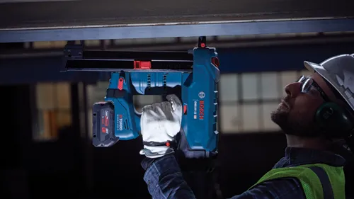 Person wearing safety glasses, ear protection, and a hard hat using a Bosch GNB18V-12 HD nailer, with 18V battery.