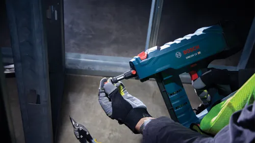 Bosch GNB18V-12 HD tool held by person wearing gloves, working on metal framing. A pair of metal shears are visible in the foreground.
