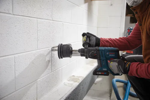 Bosch GBH18V-21 drill being used to drill into a white cinder block wall by a person wearing gloves and a respirator mask.