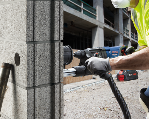 Bosch GBH18V-26D Bulldog rotary hammer drill, person wearing gloves holds it against a concrete block, the Core18V 8.0 battery is visible.