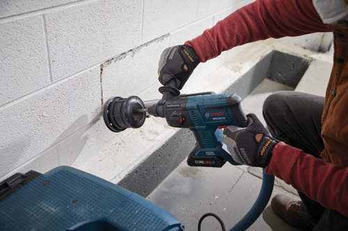 Bosch GBH18V-21 cordless Boschhammer with attached dust collection system, being used to drill into a white cinder block wall by a person wearing red long-sleeved shirt, black pants, brown jacket and work gloves.