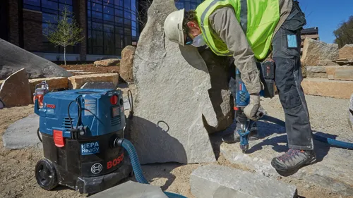 Person wearing a hard hat, face mask, safety vest, and work pants drills a rock. A Bosch vacuum cleaner "HEPA Ready" is to the left.