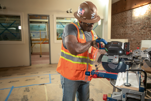A Black man wearing a safety helmet with a US flag sticker, safety glasses, and an orange safety vest, using a Bosch saw.