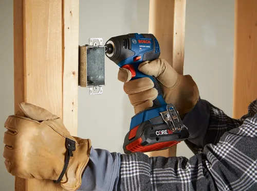 Bosch GDR18V-1800C cordless impact driver being used to attach a metal electrical box to wooden studs, wearing work gloves and a plaid shirt.