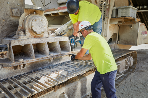 Two people wear safety gear and use a power tool near a piece of heavy machinery, one holding a Bosch CORE18V power tool.