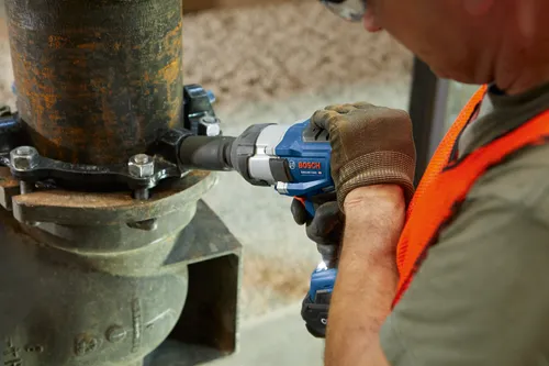 Person wearing gloves and safety glasses using a Bosch GDS18V-740C HD impact wrench on pipe hardware.