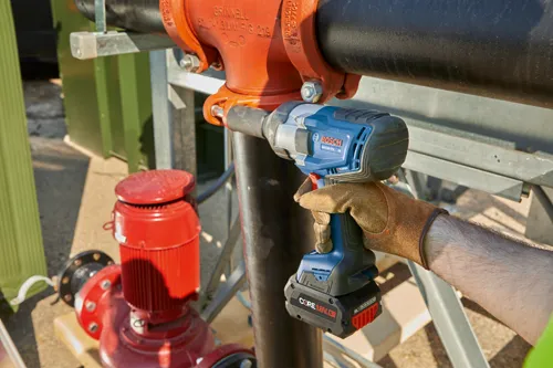Bosch GDS18V-770 impact wrench is held by a person wearing a leather glove, working on a pipe, with a red pump and other infrastructure in the background.
