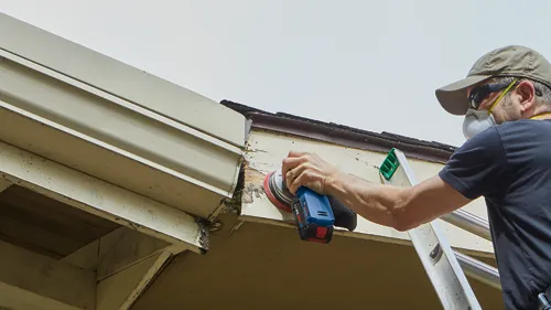 A person wearing sunglasses, a hat and a mask, uses a Bosch CORE18V sander to remove paint from a building eave while standing on a ladder.