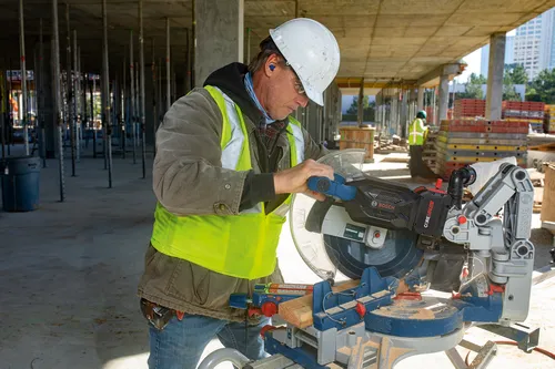 A person wearing a white hard hat, safety glasses, and earplugs is cutting wood with a Bosch power saw, while wearing a green safety vest.