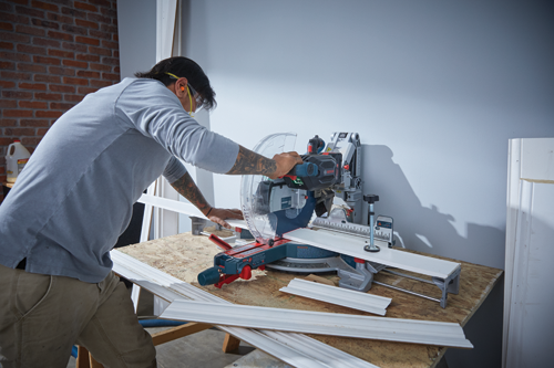 A person wearing safety glasses and ear protection uses a Bosch miter saw to cut a piece of white trim.