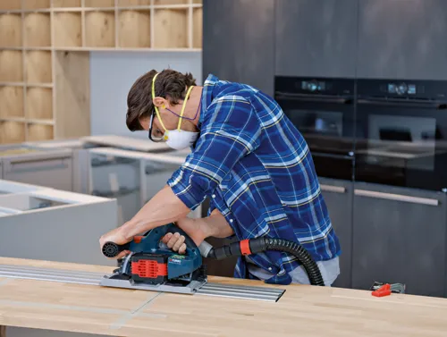 A person wearing safety glasses and a mask, in a blue plaid shirt, uses a Bosch Turbo Brushless CORE18V circular saw to cut a wooden surface.