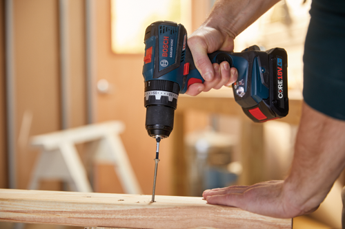 Bosch GSB18V-535C cordless drill driving a screw into a piece of wood, held by a person. CORE 18V battery is visible.