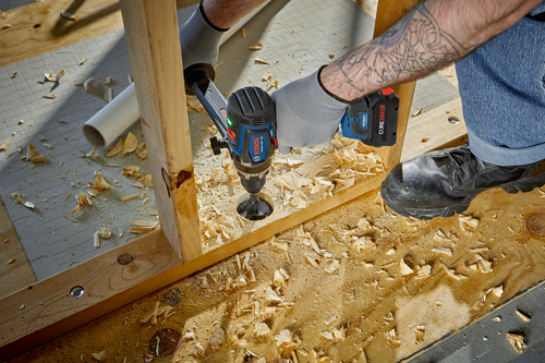 Bosch GSR18V-130C drill drilling a hole in a wooden plank, user wearing gloves and work boots.