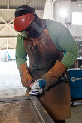 A Black person wearing protective eyewear, apron, and gloves is using an angle grinder to work on a metal frame, sparks are visible.