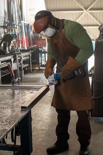 A Black person wearing protective gear, including a face shield, respirator, and gloves, is grinding metal with a grinder tool.