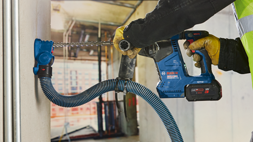 Bosch GBH18V-24C BULLDOG 18V cordless rotary hammer drill with a blue and black dust extraction system, being used by a person wearing yellow work gloves.