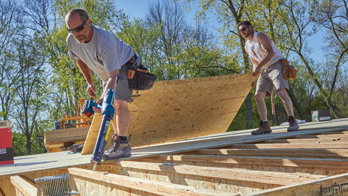 Two men working on a wooden structure, one using a caulking gun, the other carrying a large piece of wood.
