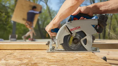 Bosch circular saw cutting wood; visible logo "BOSCH" on the saw. Another person is carrying a wooden board in the background.