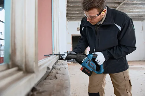 A person wearing safety glasses and ear protection is using a Bosch GSA18V-28 reciprocating saw, cutting a wood frame.