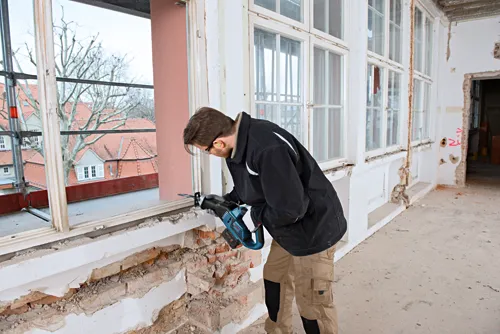 Person using a reciprocating saw to cut into the brick wall of a building.