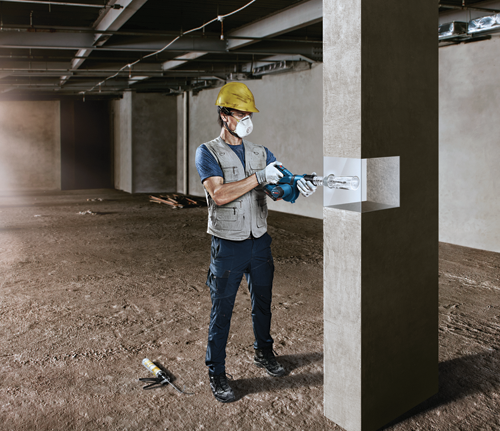 A person wearing a yellow helmet, safety glasses, and a respirator mask is operating a drill at a construction site.