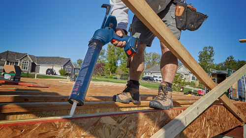 Bosch cordless caulking gun applying sealant on wooden beams, person wearing work boots and tool belt.