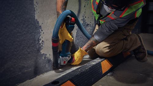 Person wearing protective gear using a grinder with a vacuum hose attached to a surface.