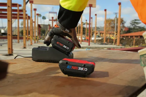 A person's gloved hand places a battery on a charging station, with another battery nearby; batteries read CORE 18V.
