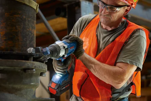 Man wearing safety glasses, a hard hat, and an orange safety vest, holding a blue and black Bosch GDS18V-770C impact wrench.