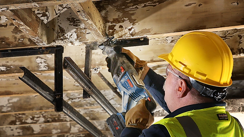 Person using a Bosch reciprocating saw, wearing a yellow hard hat and safety glasses, cutting into a wooden beam.