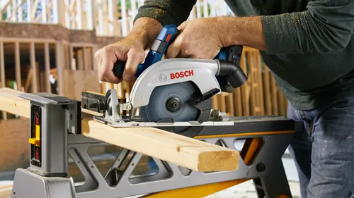 A person using a circular saw with the logo "BOSCH" to cut a wooden plank.