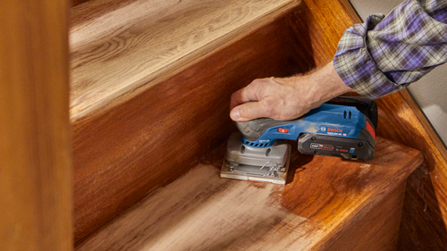 A person using a  Bosch GSS18V-40  sander on wooden stairs.