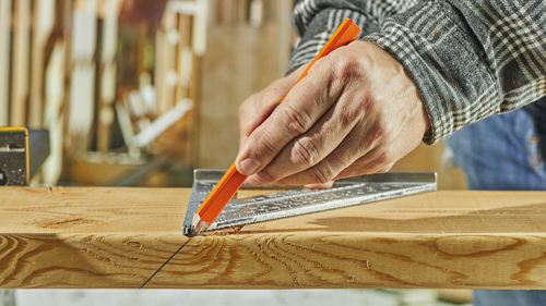 Person using an orange pencil to draw a line on a wooden plank with a metal square tool.