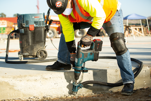 A person wearing a hard hat, face mask, and work clothes drills into concrete with a Bosch GBH18V-36C BITURBO rotary hammer drill; a Bosch HEPA vacuum cleaner is nearby, and a hose connects the vacuum to the drill.