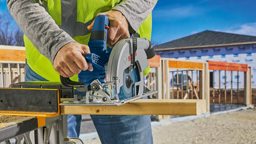 Bosch circular saw cutting wood, with a person wearing a safety vest and grey long sleeve shirt. The saw has "Bosch" visible on it. Construction site in the background.