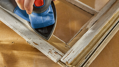 A hand sanding an old wooden window frame with a blue and black electric sander.