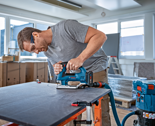 A person wearing safety glasses and ear protection, cutting a dark gray surface with a Bosch GST18V-60C jigsaw. The jigsaw is connected to a vacuum hose.
