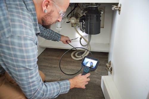 A person examines a plumbing fixture with a Bosch Professional inspection camera, inside a white cabinet.