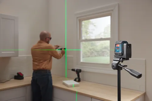Bosch laser level mounted on a tripod emitting green laser lines, a person is using the laser level to install something on the wall.
