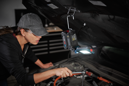 A person wearing safety glasses and a cap, working on a car engine under the hood, uses a wrench while a Bosch CORE18V 4Ah work light illuminates the area.