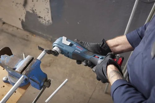 Bosch GOP18V-34 HD oscillating multi-tool cutting a metal pipe held in a vise; worker wearing black gloves.