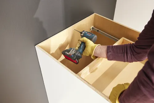 Bosch GSR12V-300FC cordless drill held by a person wearing yellow gloves, in a wooden cabinet.