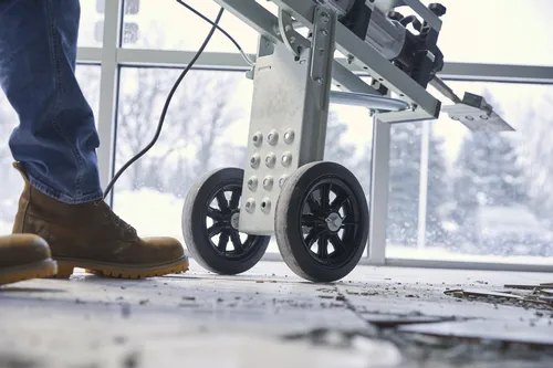 A person in blue jeans and brown work boots stands next to a wheeled apparatus with a saw blade.
