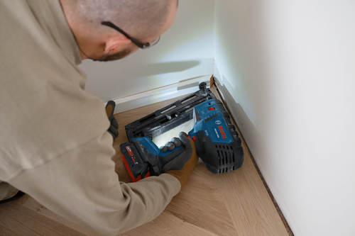 A person wearing eyeglasses and work gloves uses a Bosch GNH18V-16GA cordless nailer to install molding.