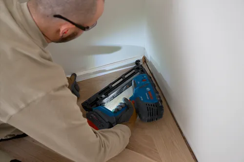 Bosch GNH18-15MA pneumatic nailer, with person wearing safety glasses and gloves, working on a floor trim.