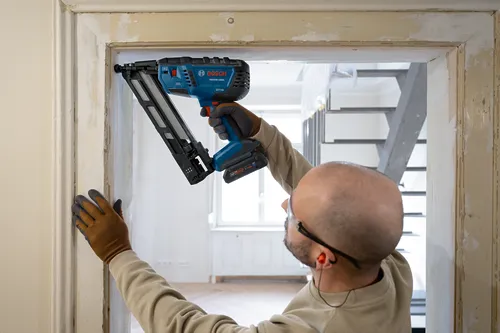 A person wearing safety glasses and earplugs uses a Bosch GNH18V-15MA nail gun.